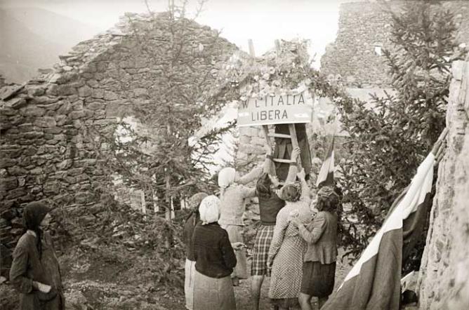 Quart (Les Trois-Villes, Avisod), 23 agosto 1945. 1° anniversario della rappresaglia nazifascista del 23 agosto 1944. Donne intente ad affiggere un cartello con la scritta «W l’Italia libera» tra le macerie. © Regione autonoma Valle d'Aosta - Foto Octave Bérard (fondo Bérard)